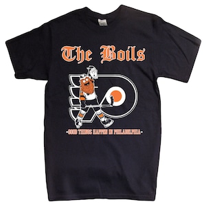 Op de afbeelding: Zwart T-shirt met de oranje tekst "The Boils" en een cartoonmascotte met een hockeystick. Het shirt bevat ook de zin "Good Things Happen in Philadelphia" in het wit.