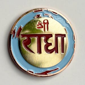 May include: A round, gold-colored metal pin with a light blue border and a red and gold design. The pin features the Hindi word "Radha" in a stylized font.