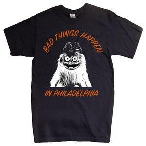 Könnte beinhalten: Schwarzes T-Shirt mit einer weißen Grafik des Maskottchens der Philadelphia Flyers, Gritty, mit einem Hockeyhelm. Der Text "Bad Things Happen in Philadelphia" ist in Orange auf dem Shirt gedruckt.