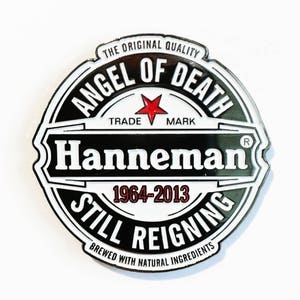 Könnte beinhalten: Ein schwarzer und weißer runder Pin mit dem Text "The Original Quality Angel of Death Trade Mark" oben herum. Der Text "Hanneman" befindet sich in einem weißen Banner in der Mitte. Der Text "1964-2013 Still Reigning" befindet sich in einem weißen Banner unten. Der Text "Brewed with Natural Ingredients" befindet sich in einer kleineren Schriftart unten herum.
