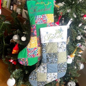 May include: Two quilted Christmas stockings hanging on a tree. The left stocking has a green cuff with the name Rachel embroidered in red and a patchwork body of green and floral fabric. The right stocking has a white cuff with the name Jacob embroidered in green and a patchwork body of blue, grey, and green paisley and floral fabric.