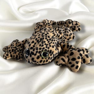 May include: A plush toy frog with a tan and black spotted pattern. The frog has large, round, black eyes with a green and gold center. The toy is lying on a white, silky fabric.