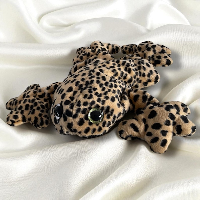 May include: A plush toy frog with a tan and black spotted pattern. The frog has large, round, black eyes with a green and gold center. The toy is lying on a white, silky fabric.