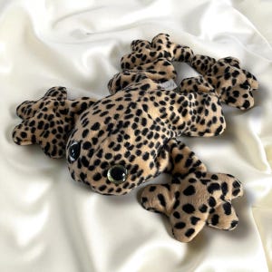 May include: A plush leopard gecko toy with a tan and black spotted pattern. The toy has large, round eyes with green centers and is lying on a white, silky surface. The gecko's body and legs are detailed.