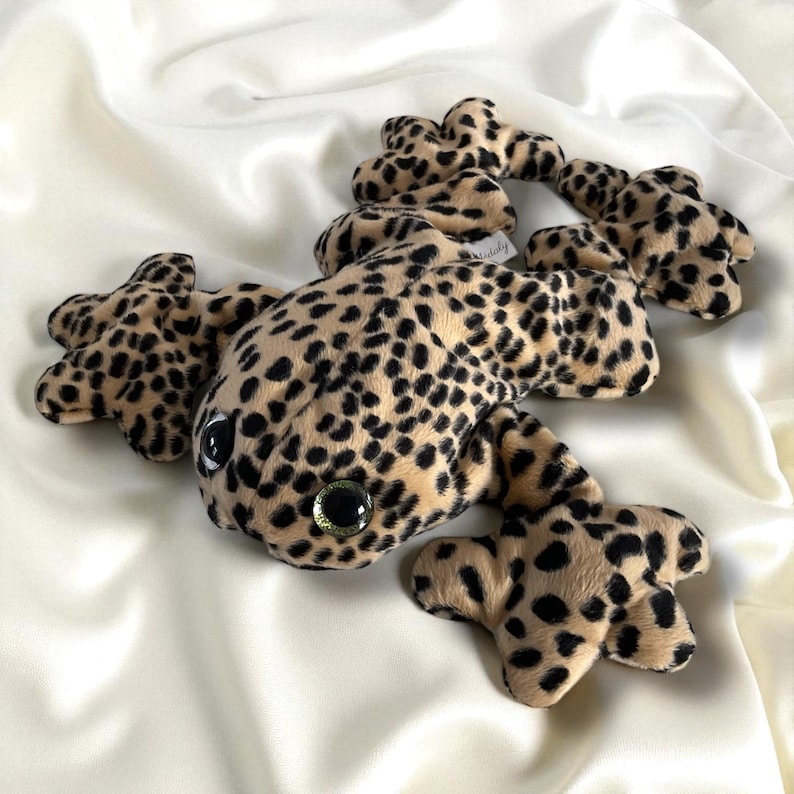 May include: A plush leopard gecko toy with a tan and black spotted pattern. The toy has large, round eyes with green centers and is lying on a white, silky surface. The gecko's body and legs are detailed.