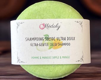 May include: A round, light green solid shampoo bar with a white label. The label reads "SHAMPOING SOLIDE ULTRA DOUX ULTRA-GENTLE SOLID SHAMPOO" and "POMME & MANGUE | APPLE & MANGO". The product is for hair care.