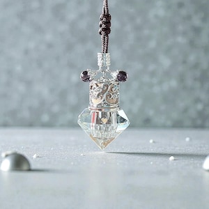 May include: An elegant perfume bottle featuring a clear, faceted crystal base and intricate silver-toned metalwork. It has a brown cord for hanging and is adorned with two dark purple beads. The design is detailed and refined.