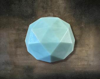 May include: A light blue, geometric-shaped object with a faceted surface. The object has a rounded, multi-sided form, with each facet catching the light. The color is a soft, muted blue-green, reminiscent of sea glass.