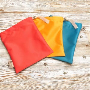 Peut inclure: Trois sacs à cordon colorés, rouge, jaune et bleu sarcelle. Chaque sac est orné d'un ruban crème et d'un charm en forme d'étoile. Les sacs sont disposés sur une surface en bois clair avec des confettis en forme d'étoile.