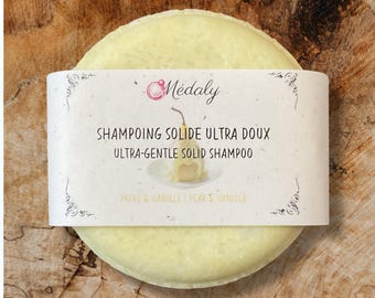 May include: A round, pale yellow solid shampoo bar with a white label. The label reads "Médaly" and "ULTRA-GENTLE SOLID SHAMPOO" with "PEAR & VANILLA" below. The bar is on a wooden surface.