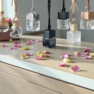 May include: A selection of hanging car air fresheners with glass bottles and wooden or black tops. The shapes include a heart, a square, and a decorative metal design. Pink and yellow flower petals are scattered on the wooden surface.