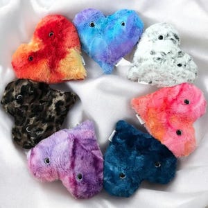 May include: Seven plush heart-shaped toys in various colours and patterns. The hearts are arranged in a circle on a white surface. The colours include red, orange, yellow, blue, purple, pink, and black.