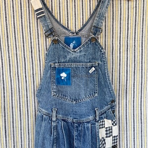 Può includere: Salopette in denim vintage con lavaggio chiaro. La pettorina presenta una piccola toppa con un motivo floreale. La salopette ha un design patchwork sulla gamba con motivi blu, neri e bianchi. Appesa a una gruccia di legno.