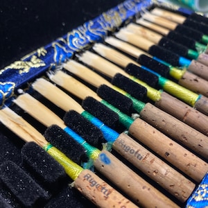 May include: A close-up of a collection of reeds for a woodwind instrument, likely a clarinet or oboe, neatly arranged in a blue and gold patterned case. The reeds are made of a light-colored material, with black and colorful accents.