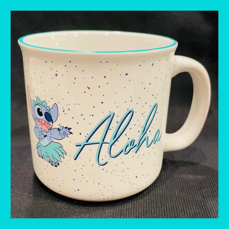 May include: White ceramic mug with a blue and green cartoon character of Stitch from Lilo & Stitch. The character is wearing a grass skirt and holding a flower. The mug has a blue rim and a speckled white background with the word "Aloha" written in blue cursive.
