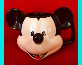 May include: A ceramic Mickey Mouse mug with black ears, a black nose, and white eyes. The mug is a light beige color and has a red tongue.