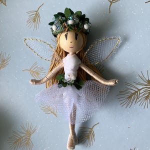 Könnte beinhalten: Eine handgefertigte Feenpuppe mit einem weißen Kleid, grünen Blättern und einer goldenen Glitzerkrone. Die Fee hat blondes Garnhaar und einen weißen Tüllrock. Die Puppe befindet sich auf einem blauen Hintergrund mit goldenen Kiefernnadeln.