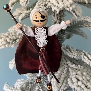 Puede incluir: Un adorno navide&ntilde;o hecho a mano que representa a un personaje caprichoso con barba blanca, parche negro en el ojo y capa color burdeos. El personaje sostiene un bast&oacute;n negro coronado con un orbe de color joya.