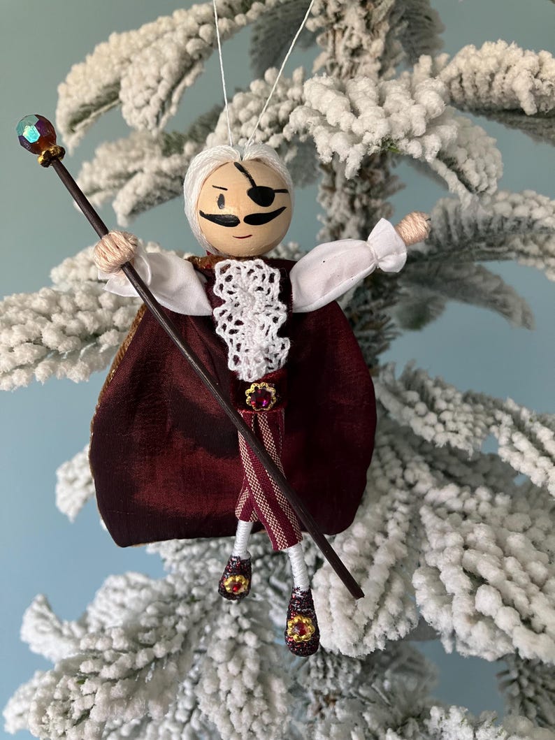Puede incluir: Un adorno navide&ntilde;o hecho a mano que representa a un personaje caprichoso con barba blanca, parche negro en el ojo y capa color burdeos. El personaje sostiene un bast&oacute;n negro coronado con un orbe de color joya.