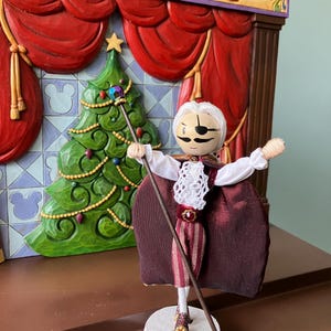 Puede incluir: Una mu&ntilde;eca de madera vestida de Cascanueces, con una capa de terciopelo burdeos, una camisa blanca y pantalones a rayas. La mu&ntilde;eca sostiene un bast&oacute;n con una estrella en la parte superior y est&aacute; de pie sobre una base redonda de madera. La mu&ntilde;eca est&aacute; frente a un escenario de madera con cortinas rojas y un &aacute;rbol de Navidad.