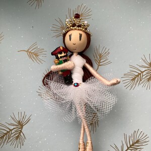 Pode incluir: Uma boneca feita à mão vestida de bailarina, com um tutu branco e uma coroa dourada com joias vermelhas. Ela está segurando um soldado de brinquedo em seus braços.