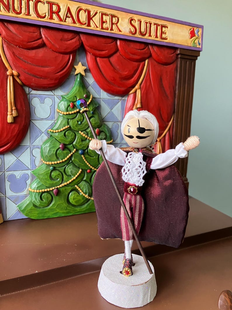 Puede incluir: Una mu&ntilde;eca de madera vestida de Cascanueces, con una capa de terciopelo burdeos, una camisa blanca y pantalones a rayas. La mu&ntilde;eca sostiene un bast&oacute;n con una estrella en la parte superior y est&aacute; de pie sobre una base redonda de madera. La mu&ntilde;eca est&aacute; frente a un escenario de madera con cortinas rojas y un &aacute;rbol de Navidad.