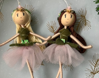 May include: Two handmade doll ornaments with green velvet dresses, pink tulle skirts, and green glitter shoes. The dolls have brown and blonde yarn hair and are wearing floral headbands with gold accents.