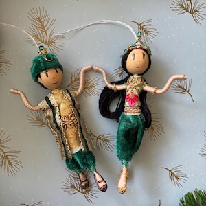 May include: Two decorative hanging ornaments featuring figures with wooden bead heads and painted faces. One figure wears a green hat and gold tunic with green pants. The other has long black hair, a beaded crown, and a green and pink outfit.