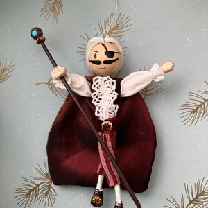Puede incluir: Un adorno de mu&ntilde;eca hecho a mano con una cara blanca, bigote negro y un parche en el ojo. La mu&ntilde;eca lleva un vestido a rayas rojas y blancas, una capa roja y una corona. La mu&ntilde;eca sostiene un bast&oacute;n de madera con una gema azul en la parte superior.