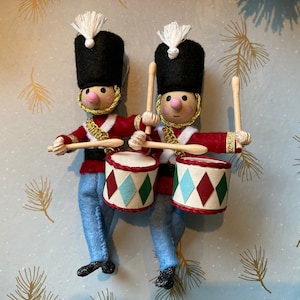 Könnte beinhalten: Zwei Filz-Spielzeugsoldaten in roten und blauen Uniformen mit schwarzen Hüten und goldenen Verzierungen, die mit Holzstöcken auf Trommeln spielen. Die Trommeln sind mit roten, grünen und weißen Diamanten verziert.