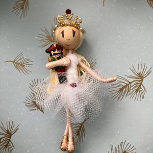 以下が含まれることがあります： 白いチュチュを着た手作りのバレリーナ人形。ミニチュアのくるみ割り人形を持っています。人形は金髪で、金の王冠があり、糸と布で作られています。背景は、金色の松の枝のデザインが施された水色です。