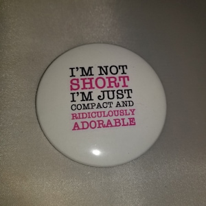 May include: White button with black text that reads "I'm not short I'm just compact and ridiculously adorable" with the words "short" and "adorable" in pink.