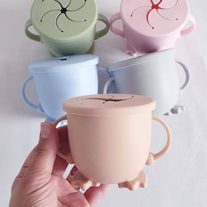 Puede incluir: Cuatro tazas de silicona con asas y una tapa en forma de estrella. Las tazas son de color verde, rosa, azul claro y beige.