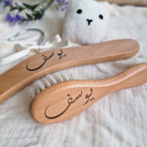 Può includere: Due spazzole per capelli in legno con il nome "Yousef" inciso in scrittura araba. Le spazzole hanno setole naturali e sono su uno sfondo di tessuto bianco.
