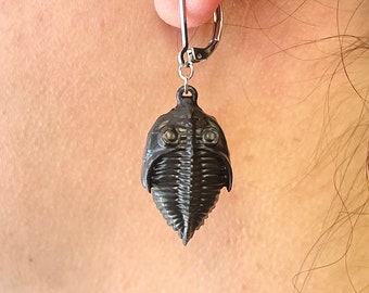May include: A single, dark gray trilobite earring with a silver-colored lever back closure. The earring features a detailed, three-dimensional design of the ancient marine arthropod. The earring is shown against a blurred background.