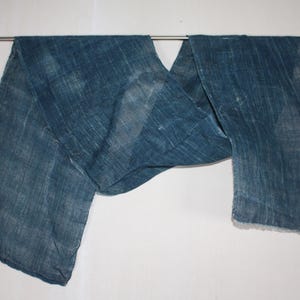 Könnte beinhalten: Ein rechteckiges Stück indigoblau gefärbter Stoff mit sichtbarer Webstruktur und ungleichmäßigen blauen Farbvariationen. Das Textil ist über eine dünne horizontale Metallstange drapiert und weist an der rechten Seite eine rohe, ausgefranste Kante auf.