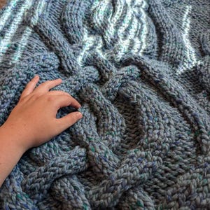 Puede incluir: Una manta de punto grueso, hecha a mano, con diseño de trenzas en tonos azules y grises. La manta presenta un diseño texturizado con hebras gruesas e entrelazadas, creando un textil acogedor y visualmente atractivo. El hilo tiene motas de color turquesa y azul.