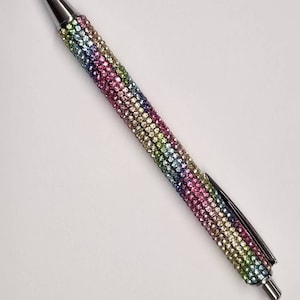 Può includere: Una penna argentata ricoperta di strass colorati. La penna presenta un motivo arcobaleno di strass rosa, verdi, blu e gialli.