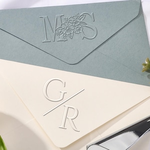 Könnte beinhalten: Zwei Umschläge, einer in Salbeigrün und einer in Creme, mit geprägten Initialen und floralen Details. Ein silberner und brauner Prägestempel liegt daneben, mit weißen Blumen im Hintergrund. Ideal für Hochzeitseinladungen oder Schreibwaren.
