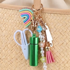 May include: A straw tote bag with a beige leather handle, adorned with a gold keychain. The keychain features a rainbow charm, a green cylinder, white scissors, and a pink tassel with floral and pearl accents.