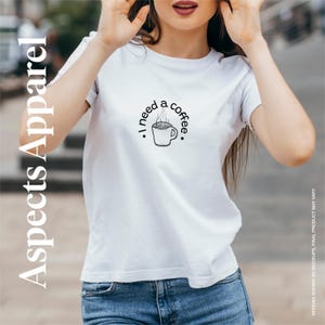 Op de afbeelding: Wit T-shirt met een zwarte afbeelding van een koffiekopje en de tekst ". I need a coffee." in een cirkelvormig ontwerp. Het shirt is van Aspects Apparel.