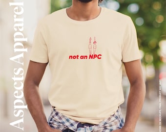 May include: A cream-coloured t-shirt featuring the red text "not an NPC" and a red graphic of two figures. The shirt is from the Aspects Apparel collection. The person is wearing a plaid shirt tied around the waist.