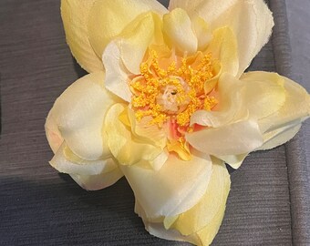 May include: A single, pale yellow silk flower with a yellow and orange center. The flower is on a dark gray background.