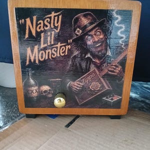 May include: A rectangular wooden box with a handle on top and a gold knob on the front. The front panel features a graphic of a grinning character in a hat, a skull, a bottle, and a cigar box labeled Zippo, with the text Nasty Lil Monster.