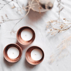 Puede incluir: Tres candelabros de cobre con velas blancas sobre una superficie blanca con flores secas en un jarrón blanco.