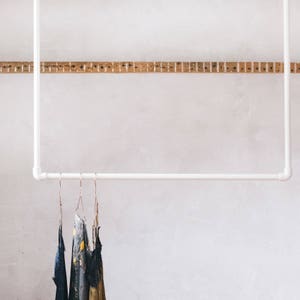 Könnte beinhalten: Drei Kleiderbügel mit verschiedenen Stoffkleidern, die an einer weißen Metallstange hängen, die an einer weißen Wand befestigt ist.
