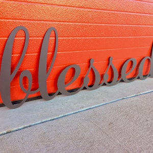 May include: A metal sign with the word "blessed" in a cursive font. The sign is dark brown and is placed on a gray concrete surface in front of a red wall.