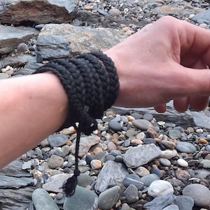 May include: A black woven bracelet wrapped around a wrist. The bracelet is made of thick, dark material, possibly rope or cord. A tassel hangs from the end. The background is a rocky beach.