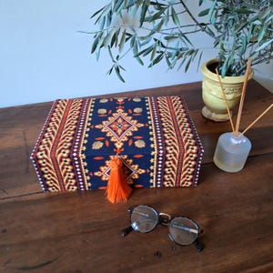 May include: A decorative box with a blue, orange, and yellow patterned fabric cover. The box has a tassel on the front and is sitting on a wooden surface.