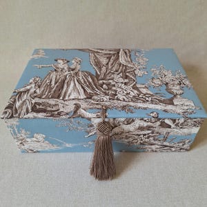 Può includere: Una scatola rettangolare con sfondo azzurro e un motivo toile de jouy marrone scuro raffigurante figure in abiti d'epoca, alberi e una fontana. Una nappa decorativa marrone abbinata pende dalla chiusura centrale sulla parte anteriore.